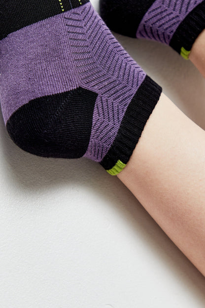 A close-up of a person's foot wearing a purple and black athletic sock with a textured chevron pattern and a small neon green accent at the ankle.