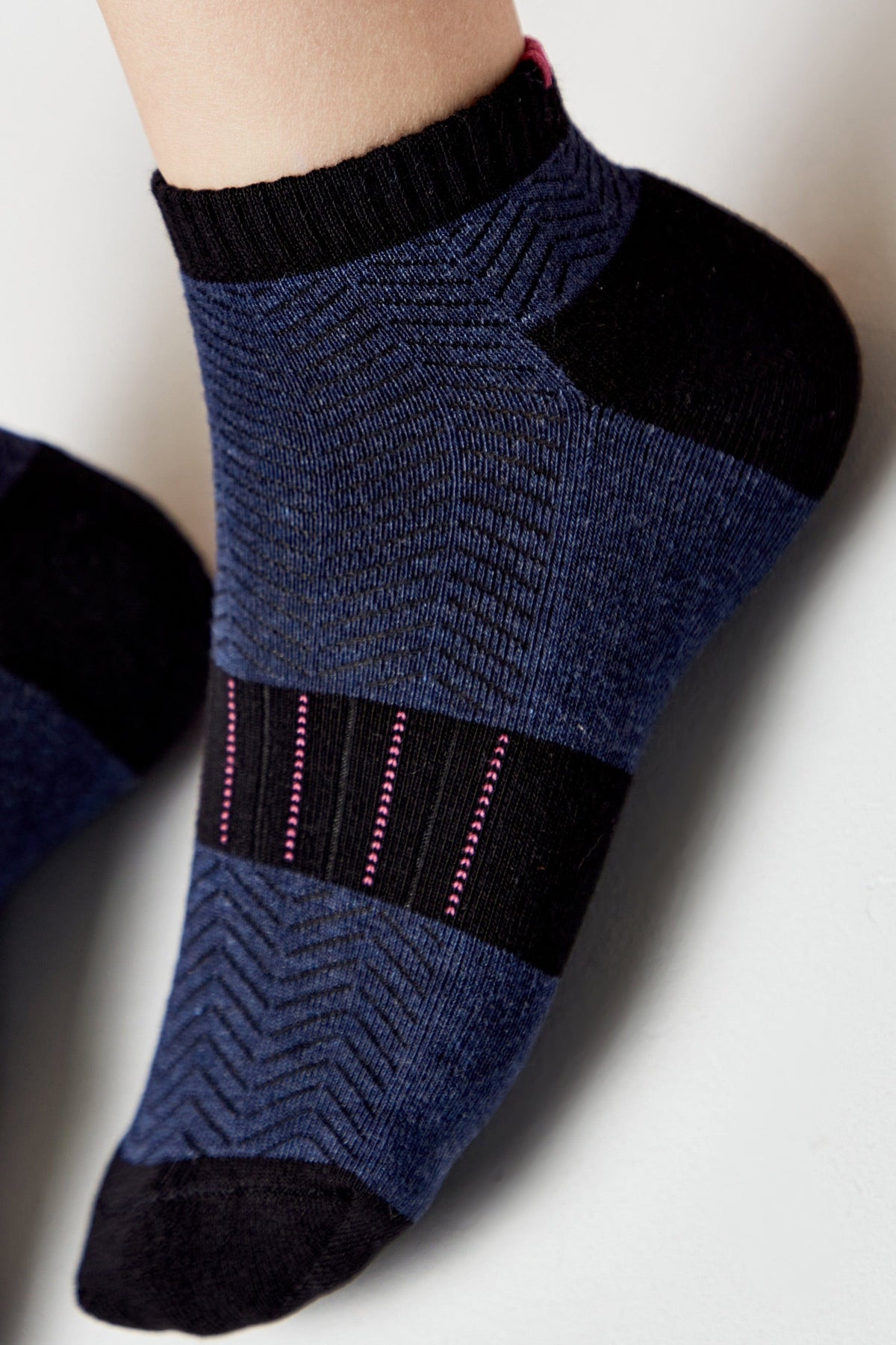 A close-up of a blue ankle sock with a black herringbone pattern, black toe and heel, and a horizontal black band across the mid-foot featuring thin vertical pink stripes.