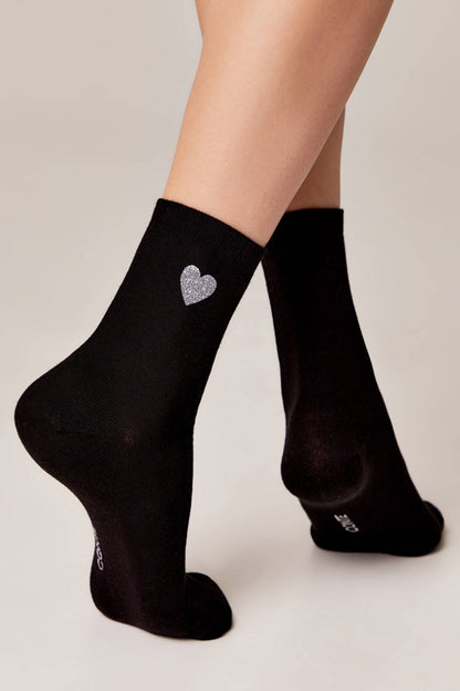A pair of black ankle socks worn on feet, featuring a small silver glitter heart design on the side of one sock.