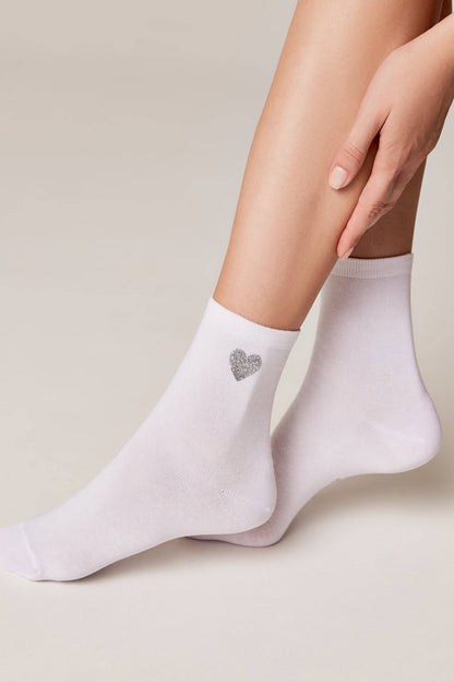 A pair of white ankle socks worn on legs, each featuring a small, silver glittery heart design on the side.