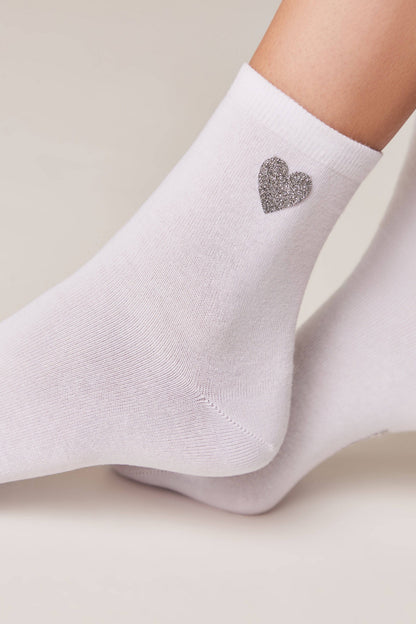 White ankle socks featuring a small, glittery silver heart detail on the side, worn on a person's feet against a light background.