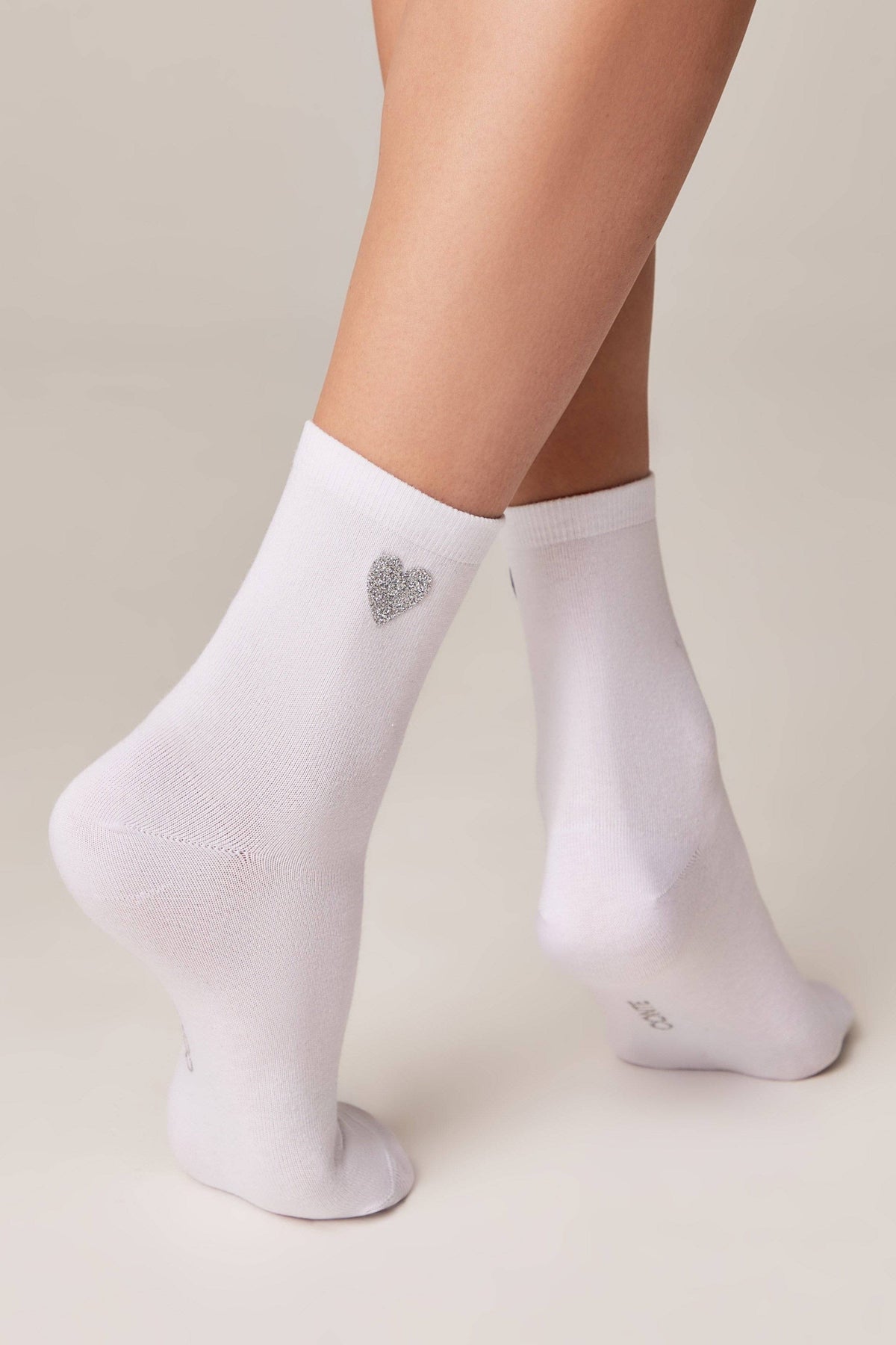 White crew socks worn on a pair of legs, featuring a small, glittery silver heart detail on the outer side of the left ankle.