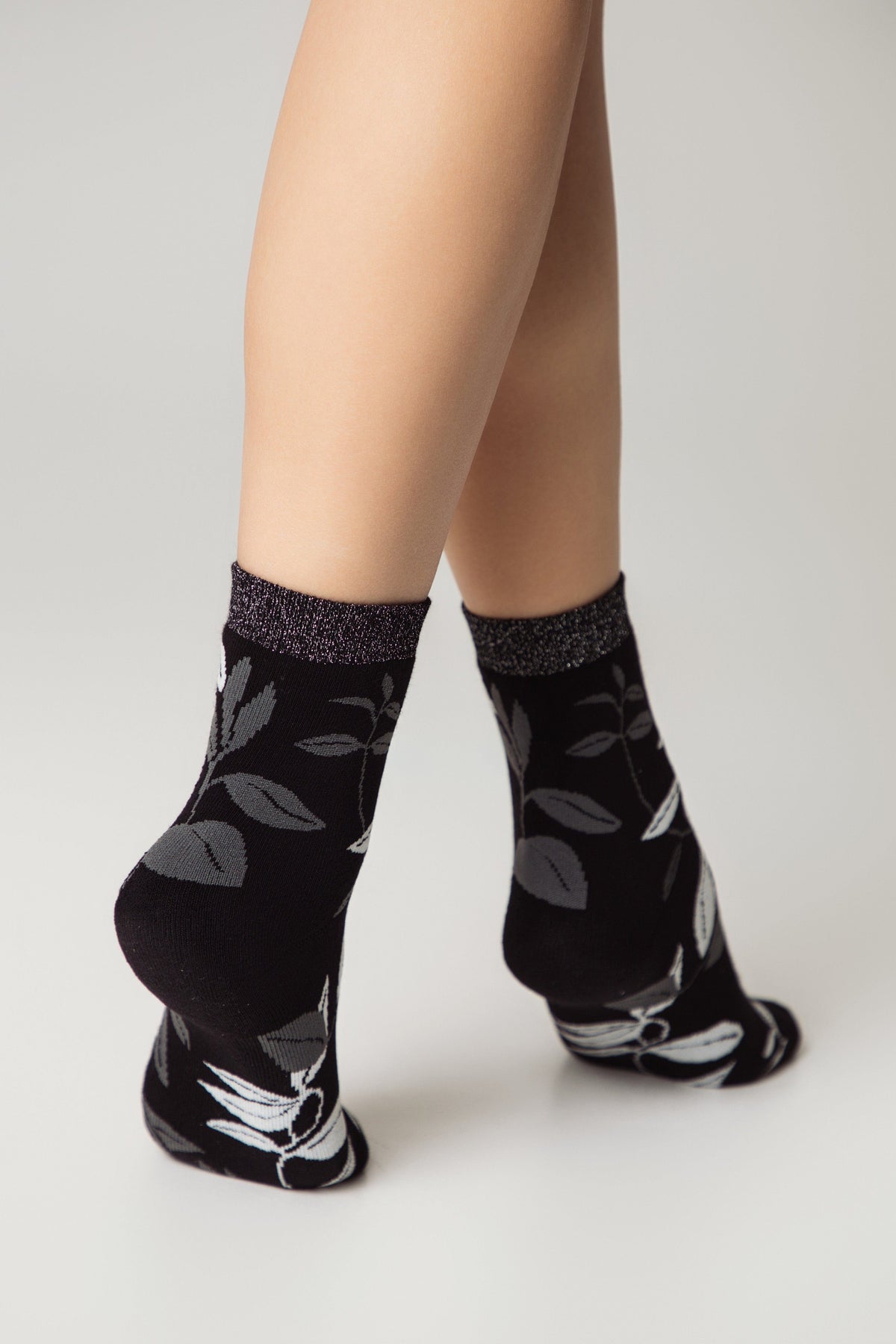 A pair of black ankle socks featuring a grey leaf pattern and a shimmering metallic grey band at the top, worn by a person standing against a plain white background.