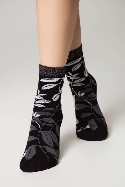 A person wears black ankle socks featuring a botanical pattern of light and dark gray leaves with a metallic silver trim at the cuff against a plain white background.