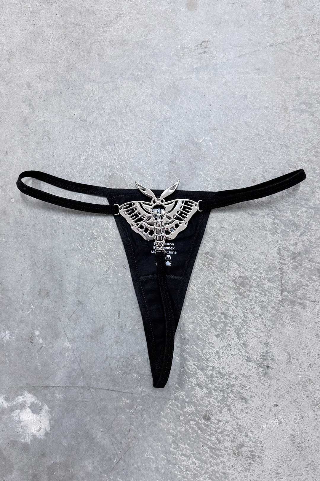 A black thong featuring a large silver-colored metal moth ornament centered on the front waistband.
