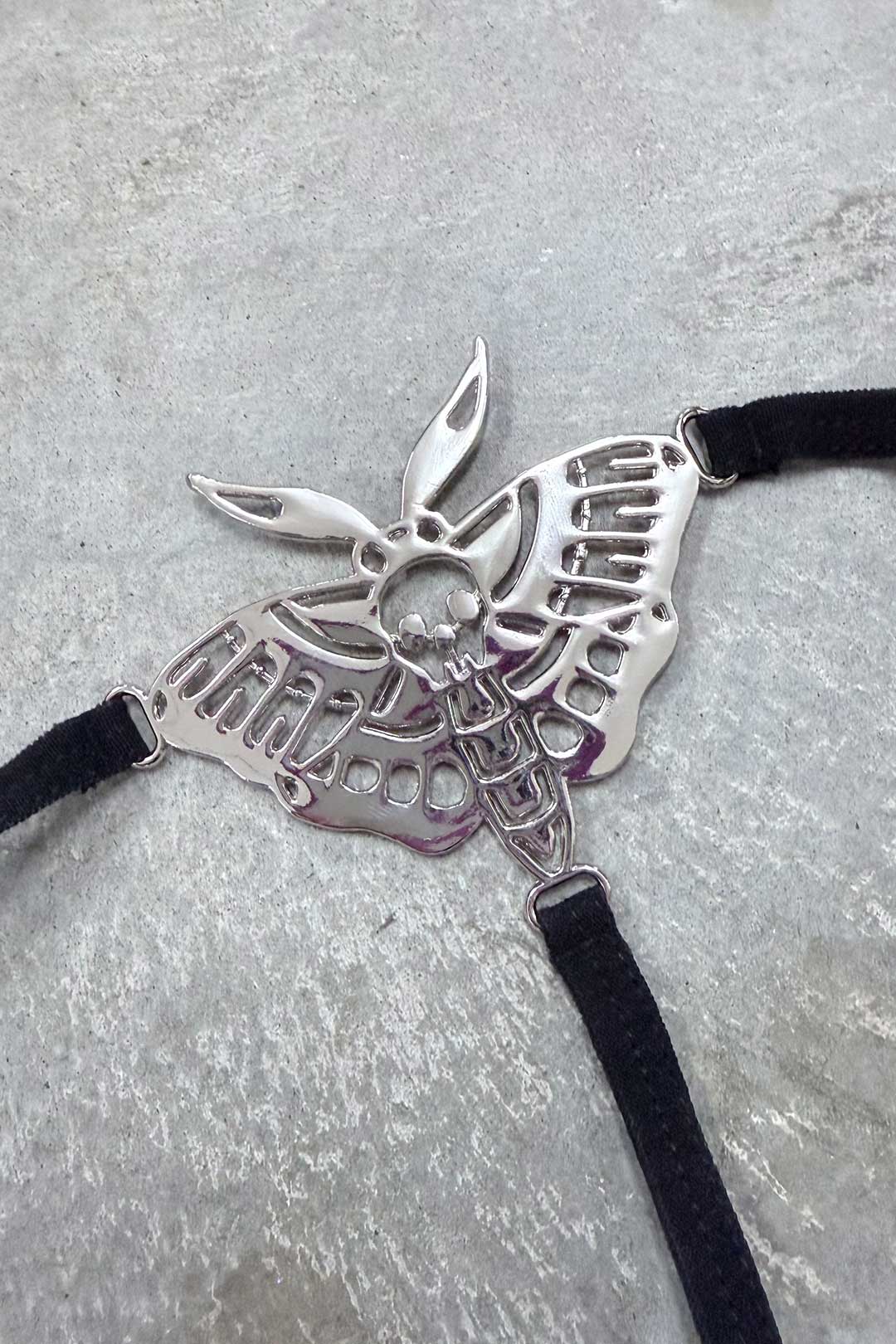 A silver-toned moth-shaped harness charm featuring a skull detail on its body, attached to black velvet-textured straps against a textured grey background.