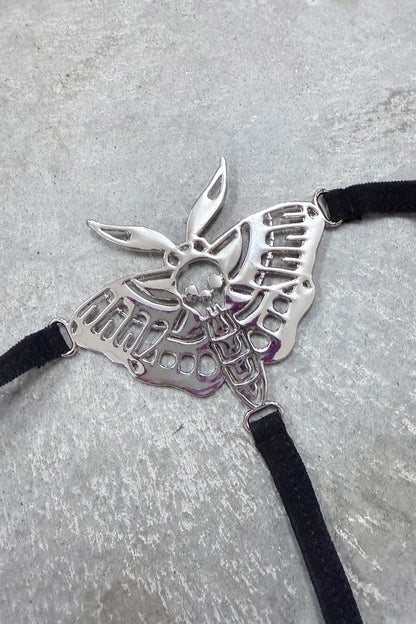 A silver-toned moth-shaped harness charm featuring a skull detail on its body, attached to black velvet-textured straps against a textured grey background.