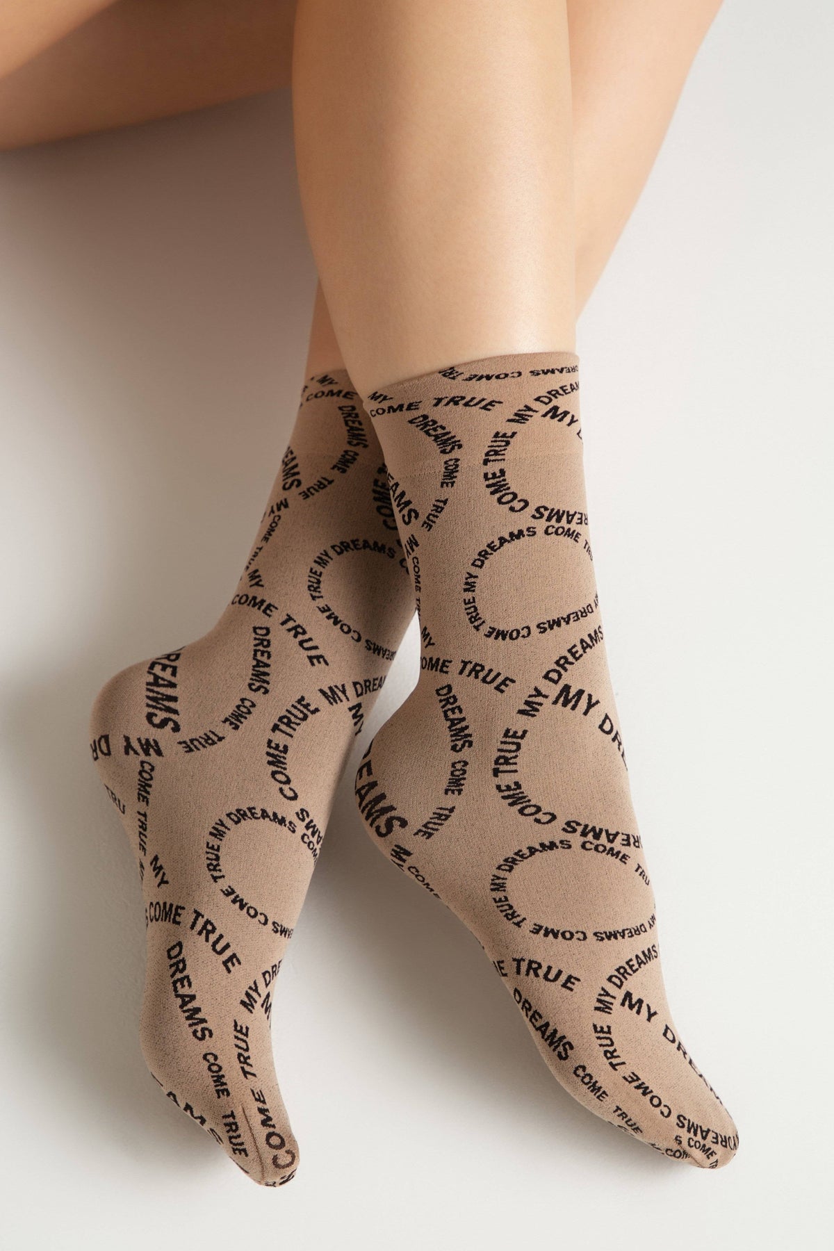 A pair of tan-colored socks featuring a repeated circular pattern of the black text MY DREAMS COME TRUE displayed on the feet and legs of a person against a white background.