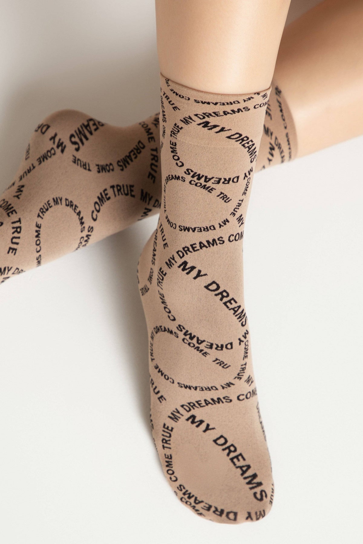 A pair of tan-colored socks featuring a repetitive, circular pattern of the black text MY DREAMS COME TRUE printed across the fabric while worn on a person's lower legs.