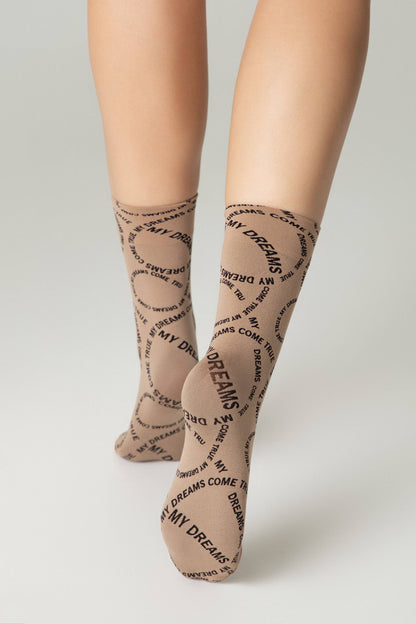 A pair of tan-colored crew socks patterned with the repeating text "MY DREAMS COME TRUE" in a circular black font, worn on a person's lower legs.