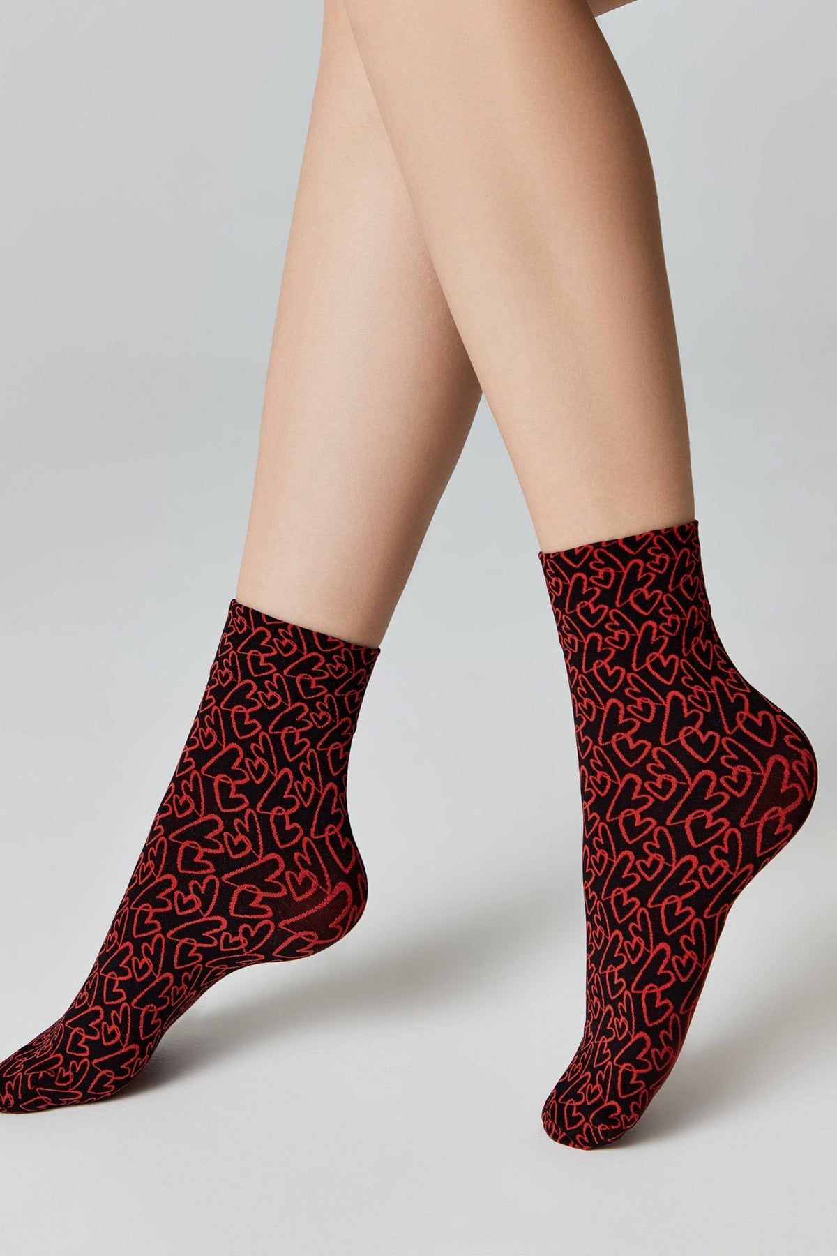 A pair of ankle socks featuring a repeating red heart pattern on a black background worn on a model's feet against a plain light gray backdrop.
