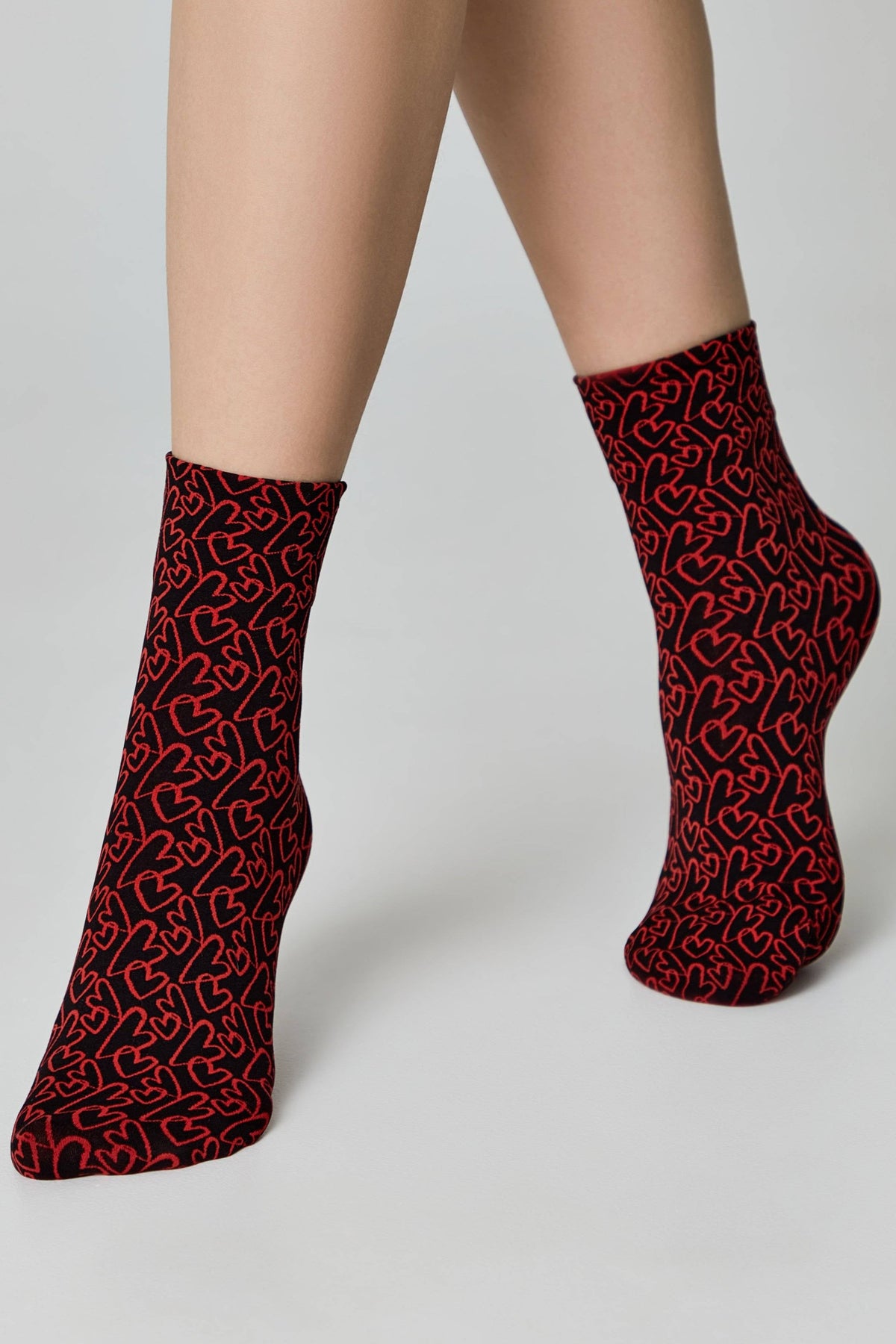 Ankle-length socks featuring a repeating pattern of red outlined hearts on a black background worn on a pair of legs against a neutral backdrop.