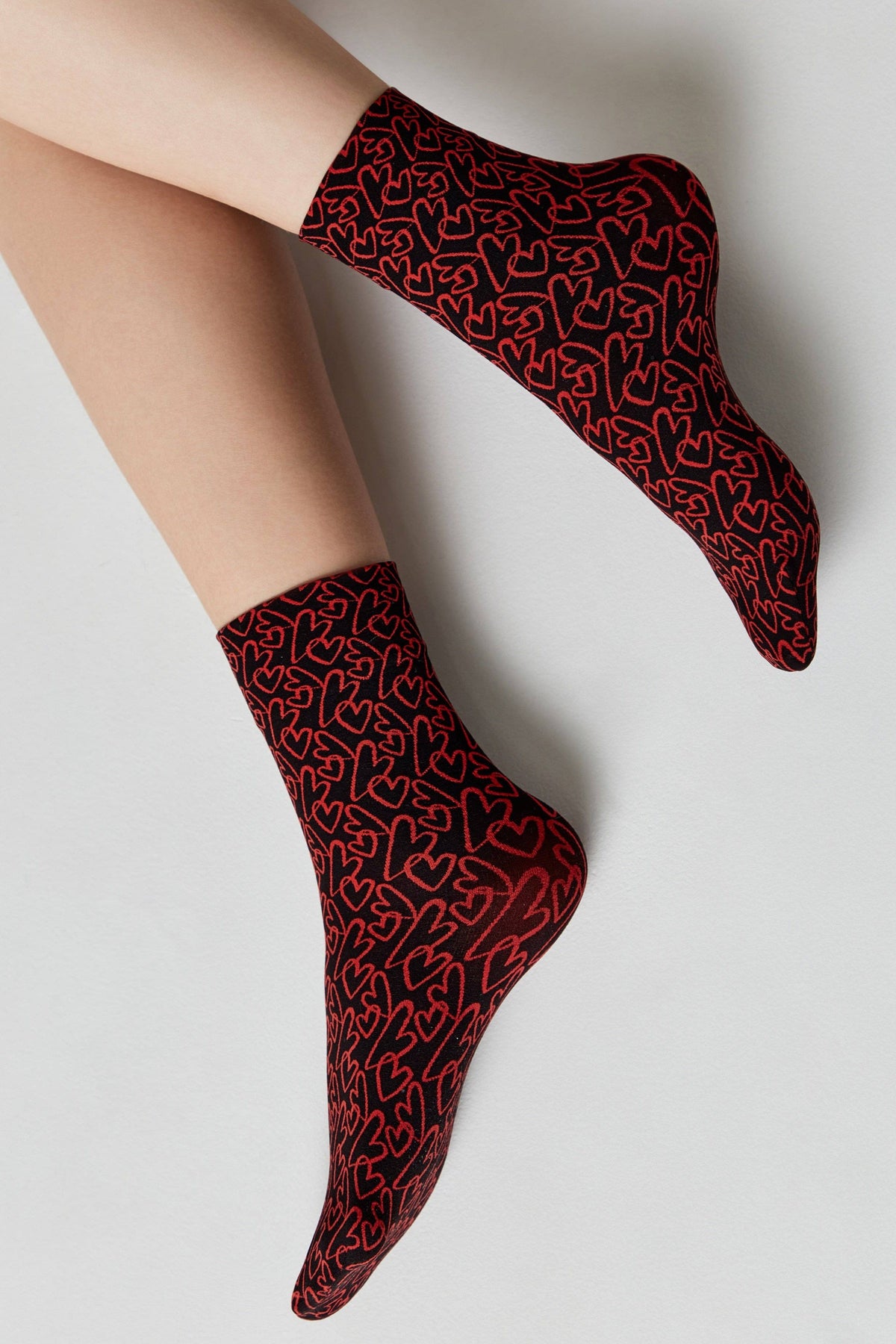 A pair of black ankle socks covered in a repeating pattern of outlined red hearts worn on a person's feet against a light grey background.