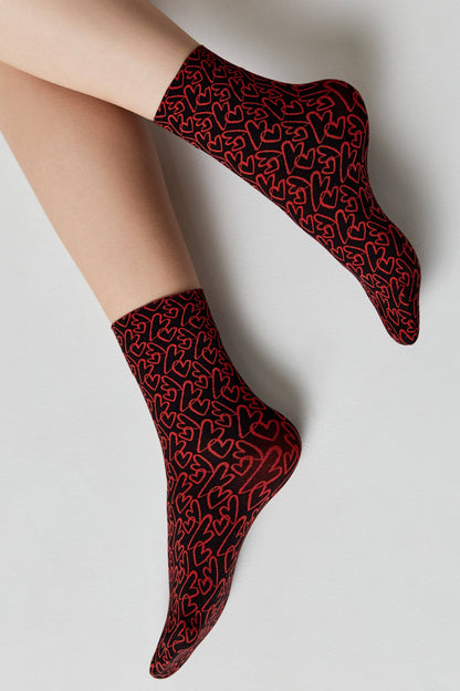 A pair of black ankle socks covered in a repeating pattern of outlined red hearts worn on a person's feet against a light grey background.