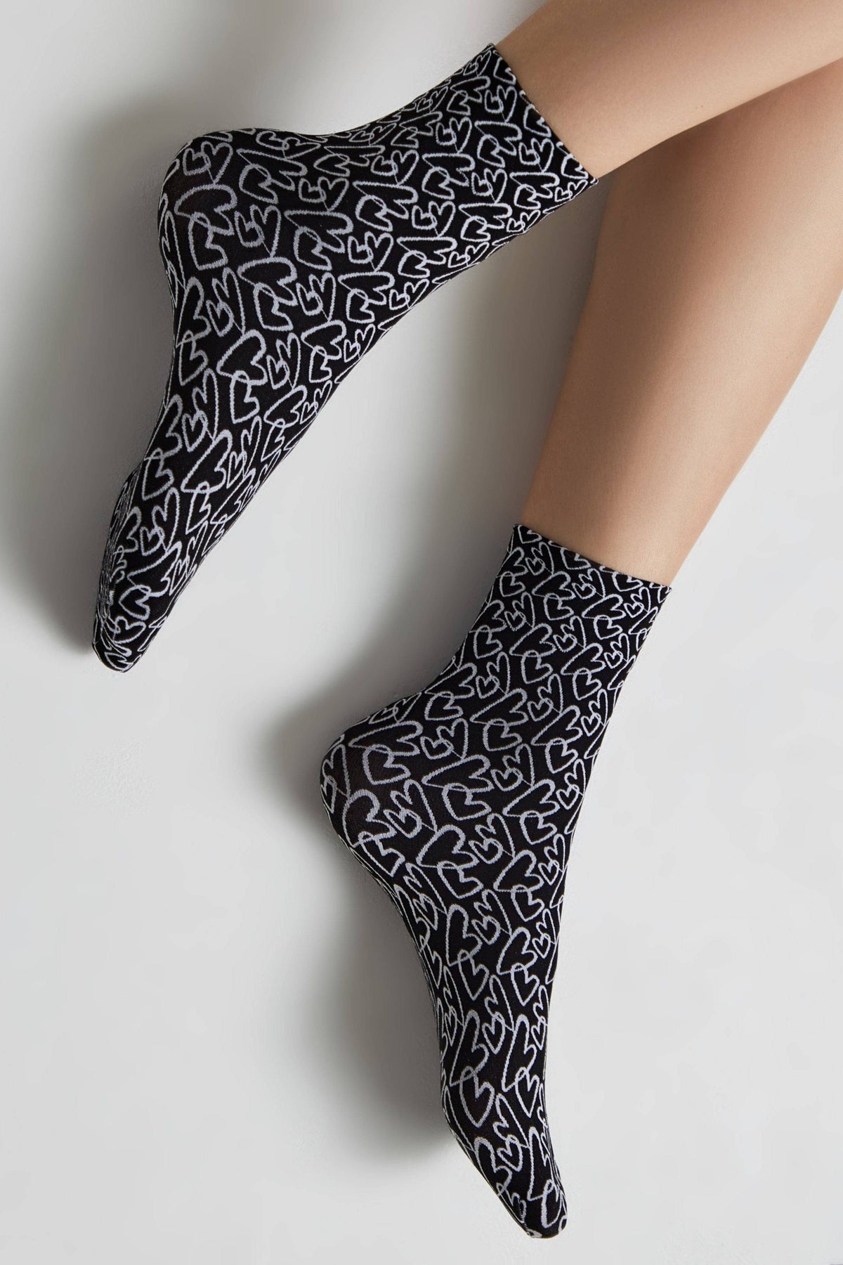 A pair of black ankle socks featuring an all-over white outline heart pattern, worn on a person's feet against a plain light background.