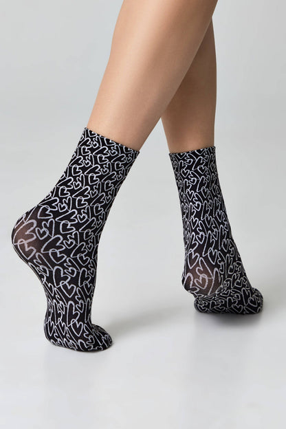 A pair of black socks patterned with white outlined heart shapes, worn on a person's feet against a plain white background.