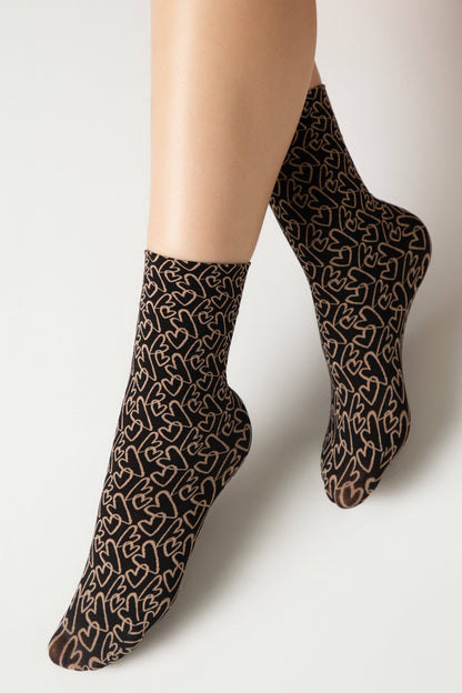 Ankle-high socks in black featuring an all-over pattern of outlined beige hearts, worn on a person's feet against a plain white background.