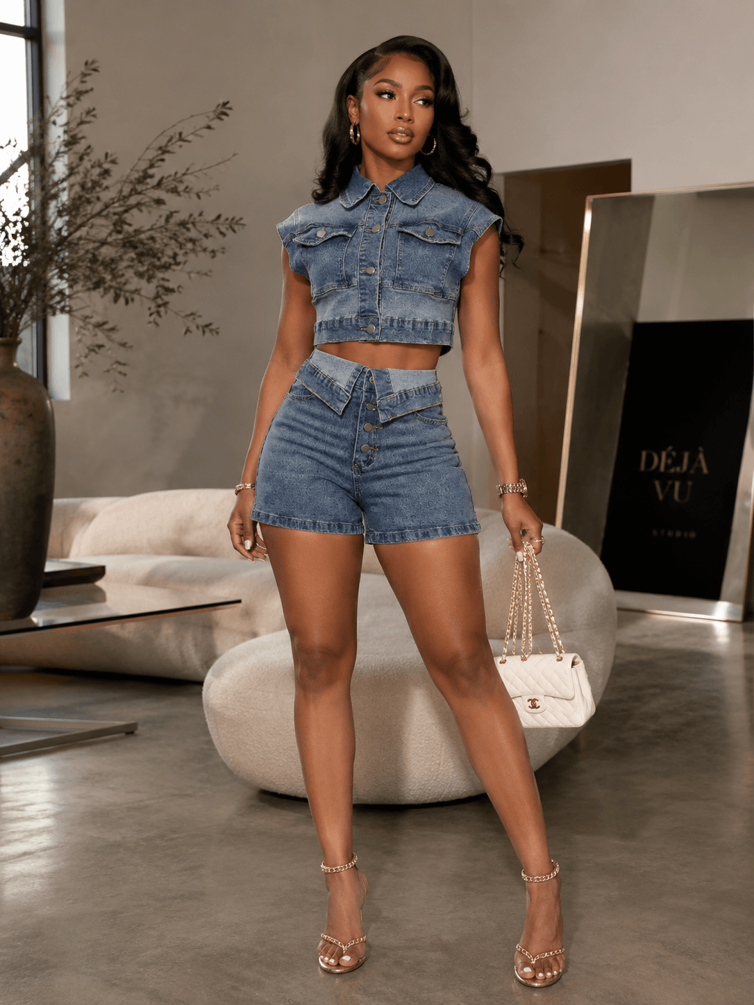 A model wearing a dark wash denim short-sleeve button-up crop top and matching high-waisted denim shorts in a modern living room setting.
