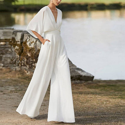 A woman stands outdoors wearing a floor-length white jumpsuit featuring a deep V-neckline, wide kimono-style sleeves, a fitted waist, and loose, wide-leg trousers.