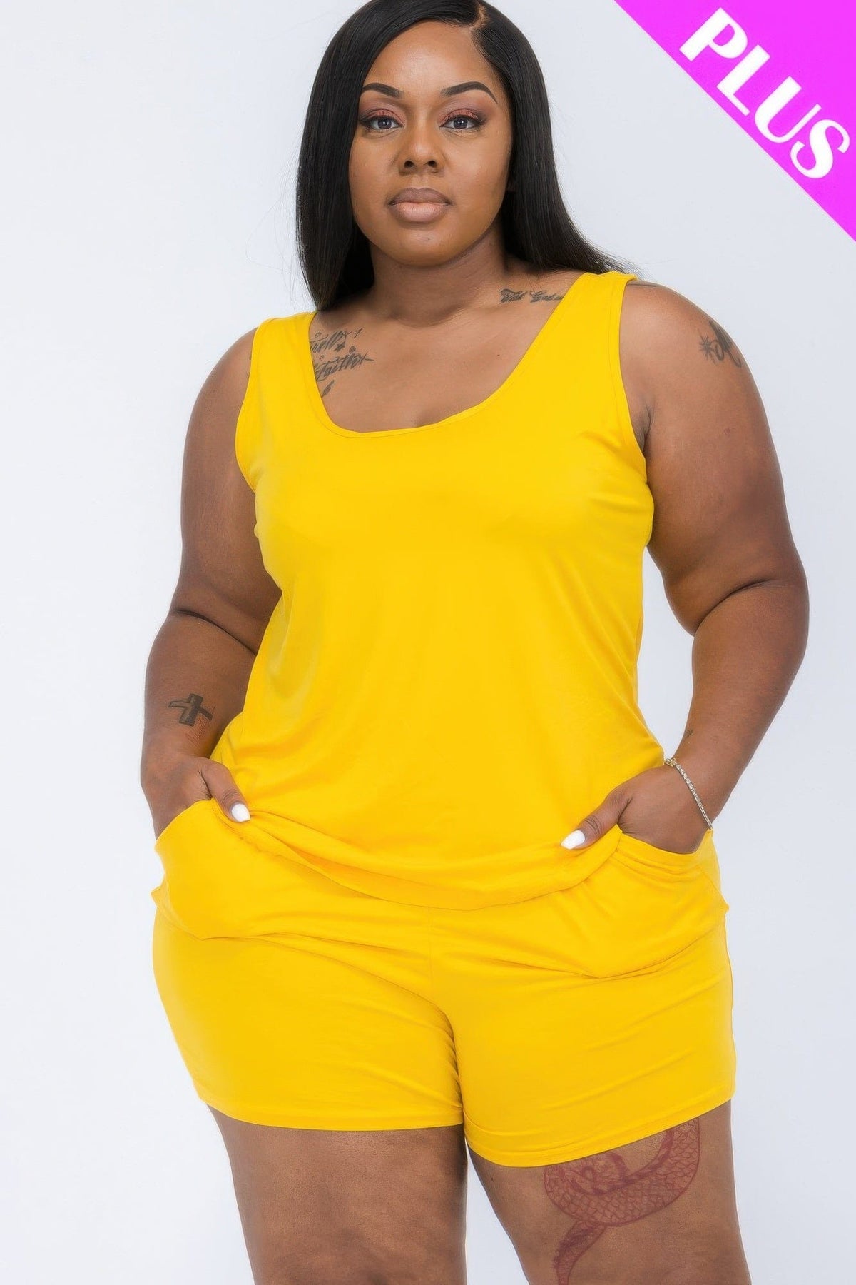 A woman wearing a solid yellow sleeveless tank top and matching shorts with pockets, standing against a white background with a bright pink plus size label in the corner.