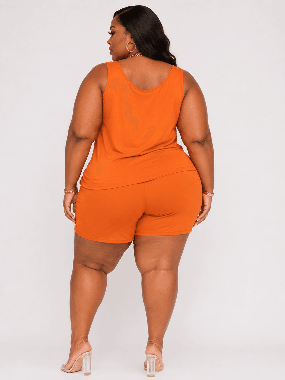 A plus-size woman stands with her back to the camera, wearing an orange sleeveless tank top and matching high-waisted shorts with clear high-heeled sandals.