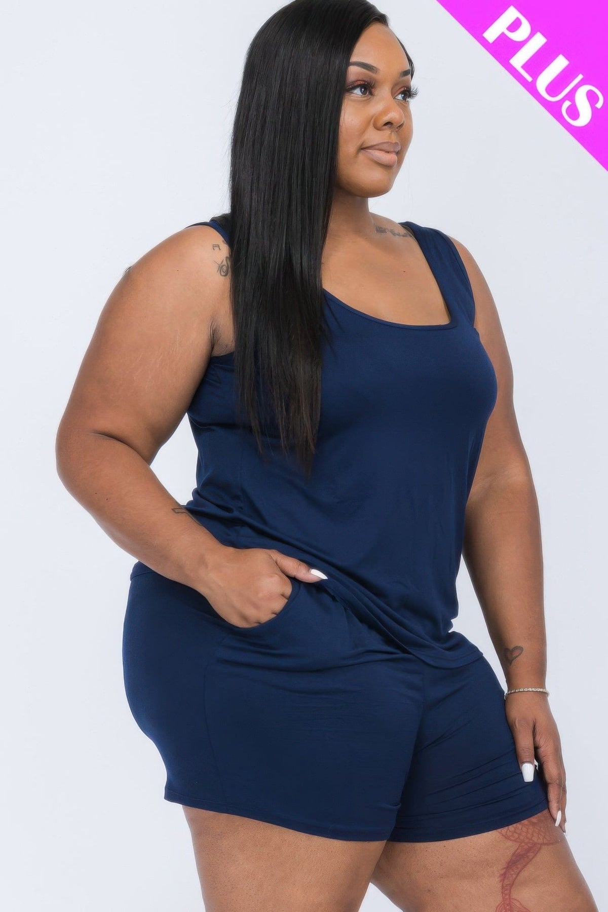 A woman stands in profile wearing a dark blue sleeveless scoop-neck tank top paired with matching shorts that feature side pockets, set against a plain white background.