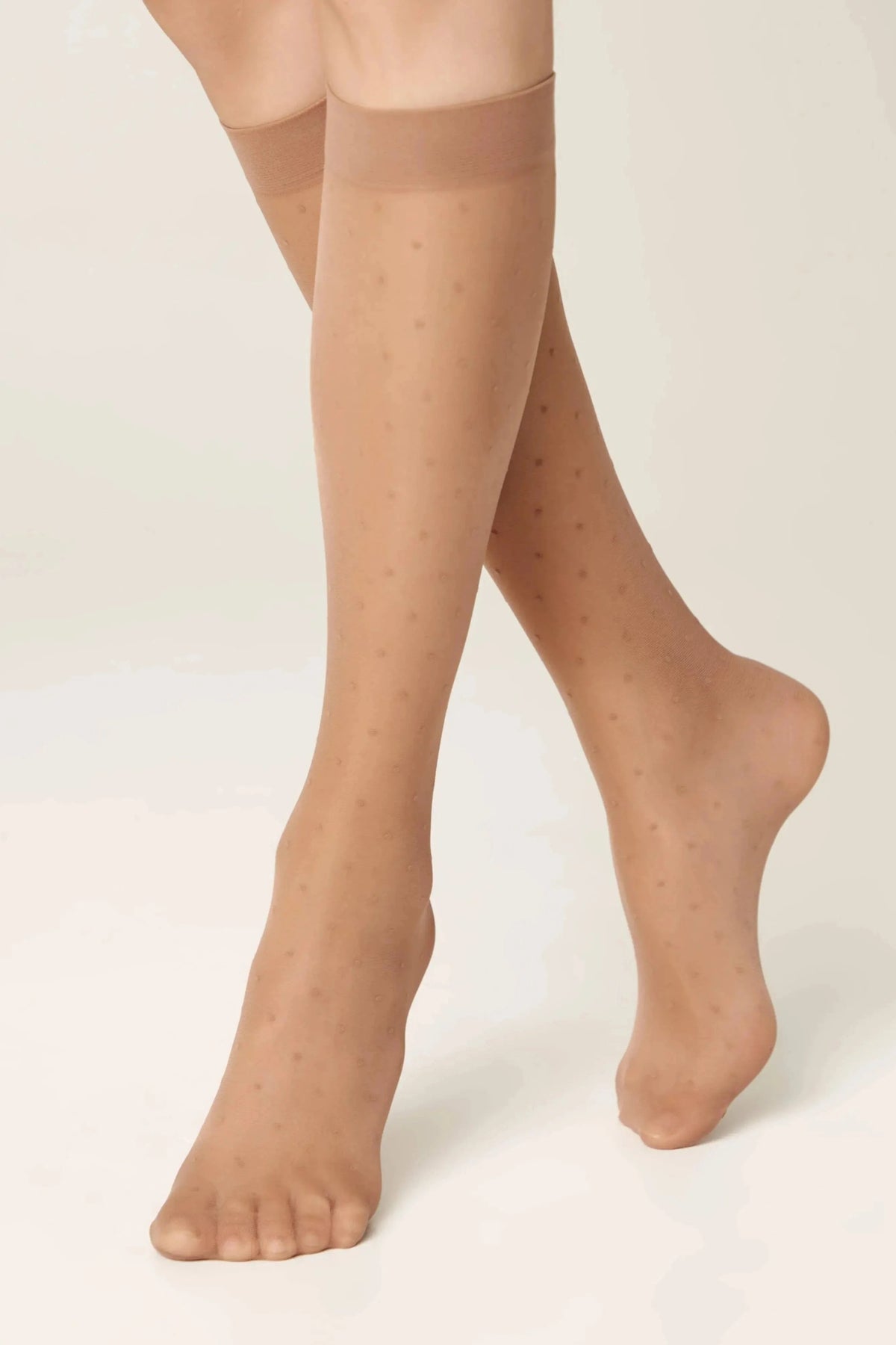 A pair of legs wearing sheer beige knee-high stockings featuring a subtle polka dot pattern against a plain light background.