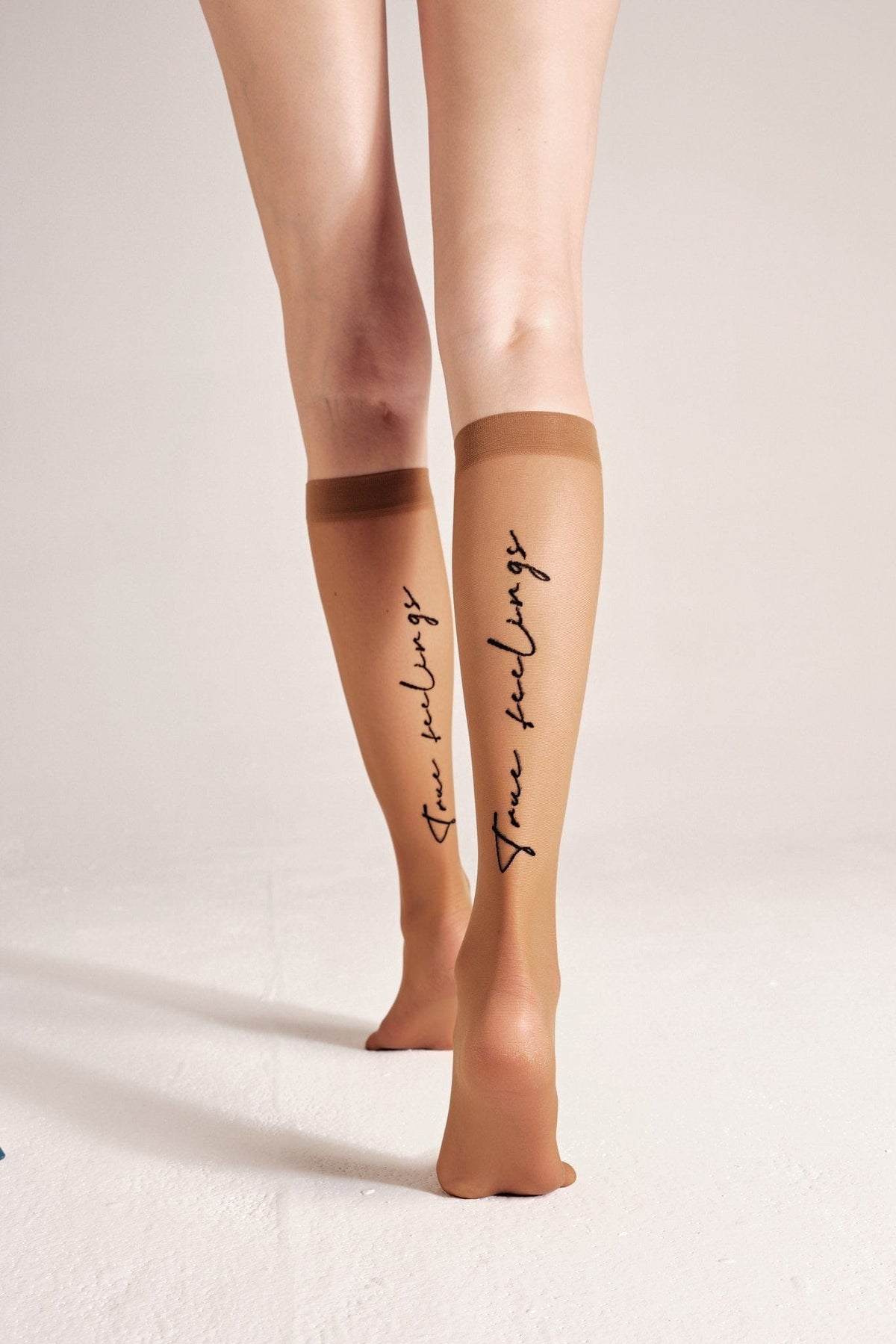 A pair of sheer tan knee-high socks worn on a person's lower legs, featuring the words 'true feelings' printed in black cursive script down the back of each calf.