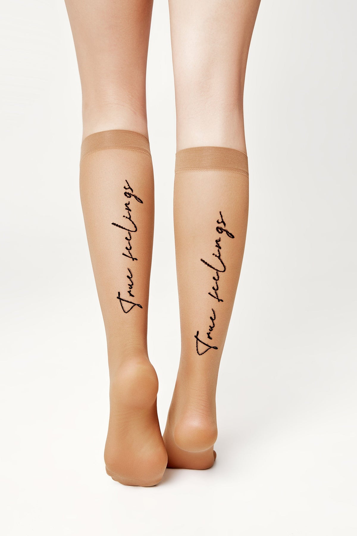 A pair of sheer beige knee-high stockings worn on the lower legs, featuring the words true feelings written in a cursive black font down the back of each calf.
