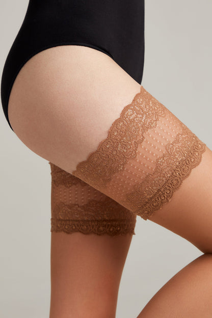 A close-up of a person wearing brown lace thigh bands on their upper legs, positioned against a plain light-colored background.