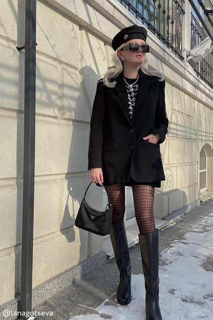 A black leather shoulder bag with a silver logo plaque held by a woman wearing a black blazer, patterned tights, and knee-high boots.