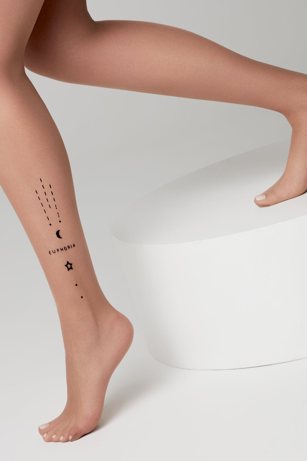 A close-up of a person's lower leg and foot wearing sheer nude tights, featuring a minimalist black temporary tattoo with a crescent moon, star, and the word EUPHORIA.