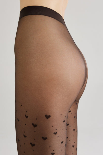 Sheer black tights featuring a repeating pattern of small dark hearts and scattered dots worn on a person's lower torso and legs against a light grey background.