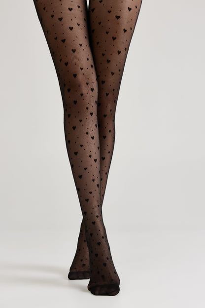 Sheer black tights decorated with a recurring small heart pattern worn on legs against a plain light gray background.