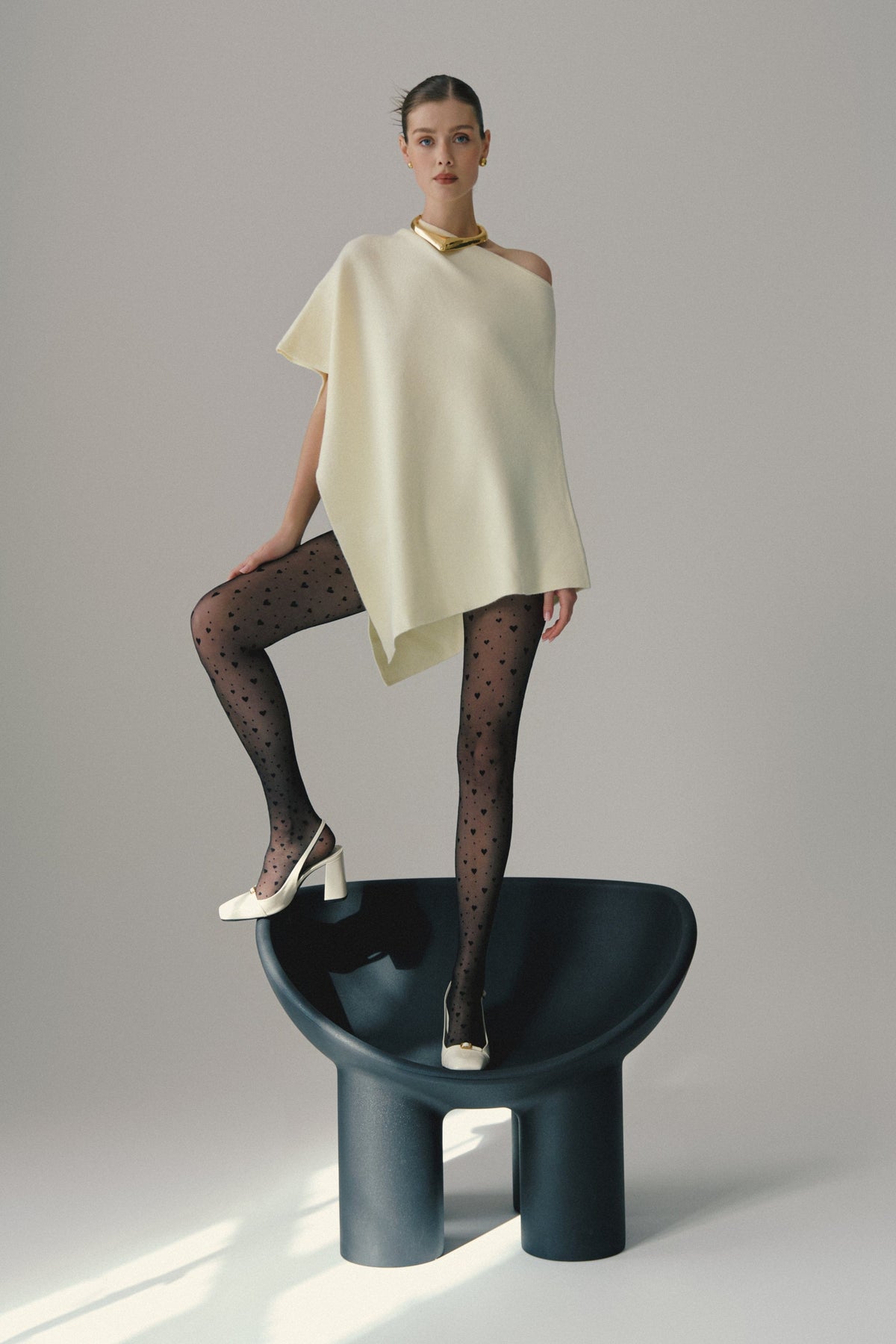 A model stands on a dark, curved chair wearing an off-white asymmetrical sleeveless top, black patterned tights with small heart shapes, and white high-heeled slingback shoes.
