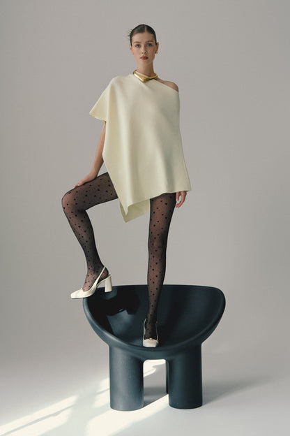 A model stands on a dark, curved chair wearing an off-white asymmetrical sleeveless top, black patterned tights with small heart shapes, and white high-heeled slingback shoes.