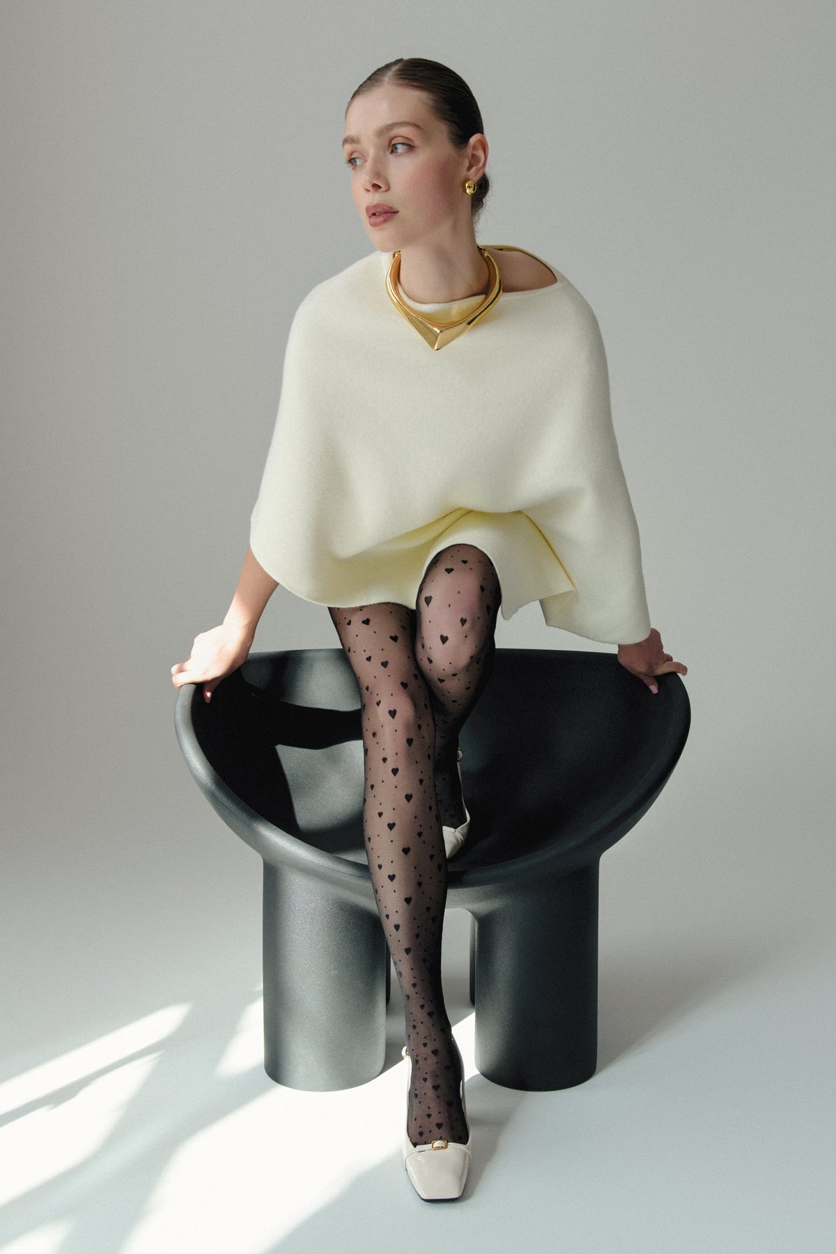 A woman sits on a black sculptural chair wearing a cream-colored poncho over black patterned tights with small heart designs and white, square-toed pumps with gold accents, complemented by a large gold choker necklace.