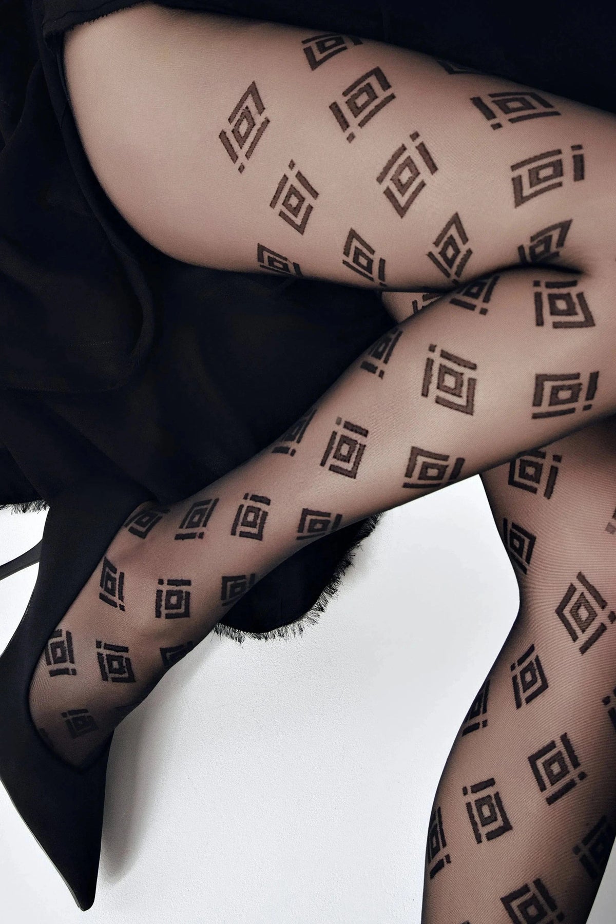 Sheer black pantyhose patterned with repeating dark geometric diamond and square symbols worn with black high-heeled shoes.