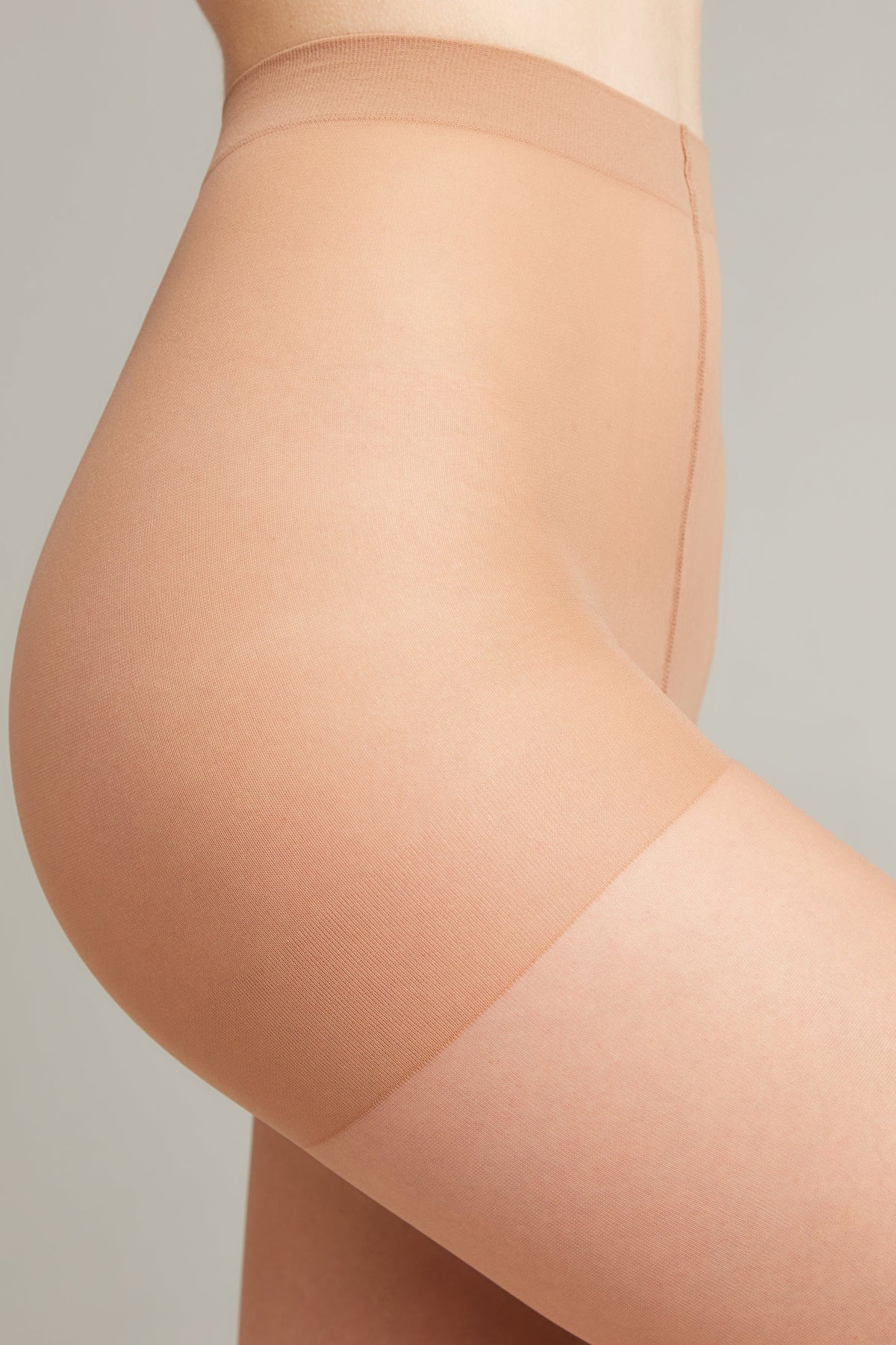 A close-up side profile of a person wearing sheer, light beige hosiery that features a reinforced waistband and upper thigh area.