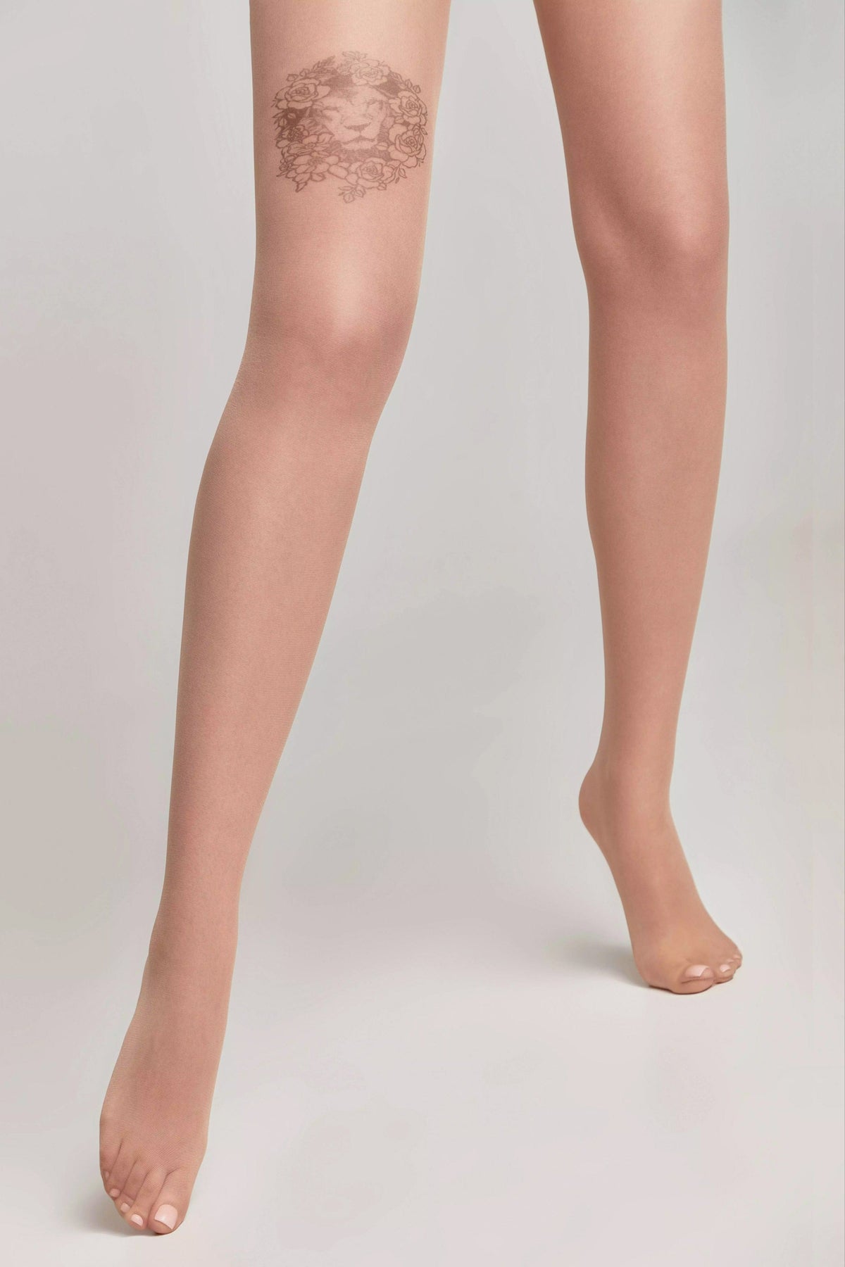 Sheer nude-colored tights worn on legs, featuring a decorative floral lion design on the upper right thigh against a plain light gray background.