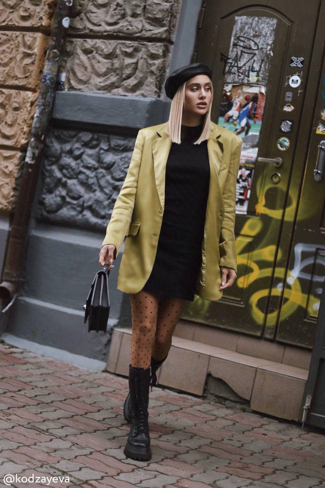 A light olive green blazer worn over a black mini dress with polka dot tights and black chunky lace-up boots, complemented by a black beret and a matching handbag while walking on a paved street.