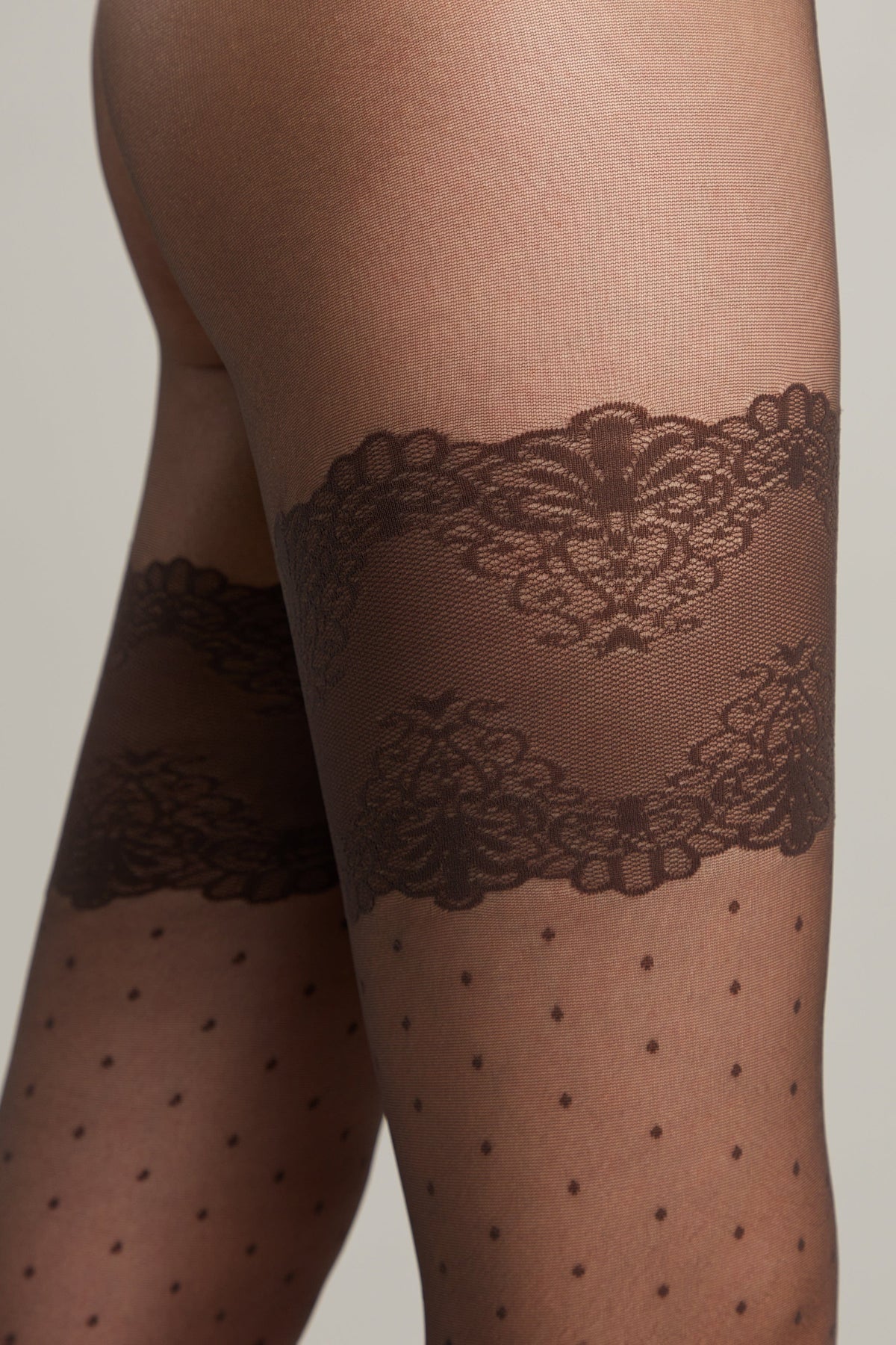 Close-up of a person wearing sheer black hosiery featuring a decorative dark brown lace garter band pattern above the knee and a subtle polka dot design on the legs.