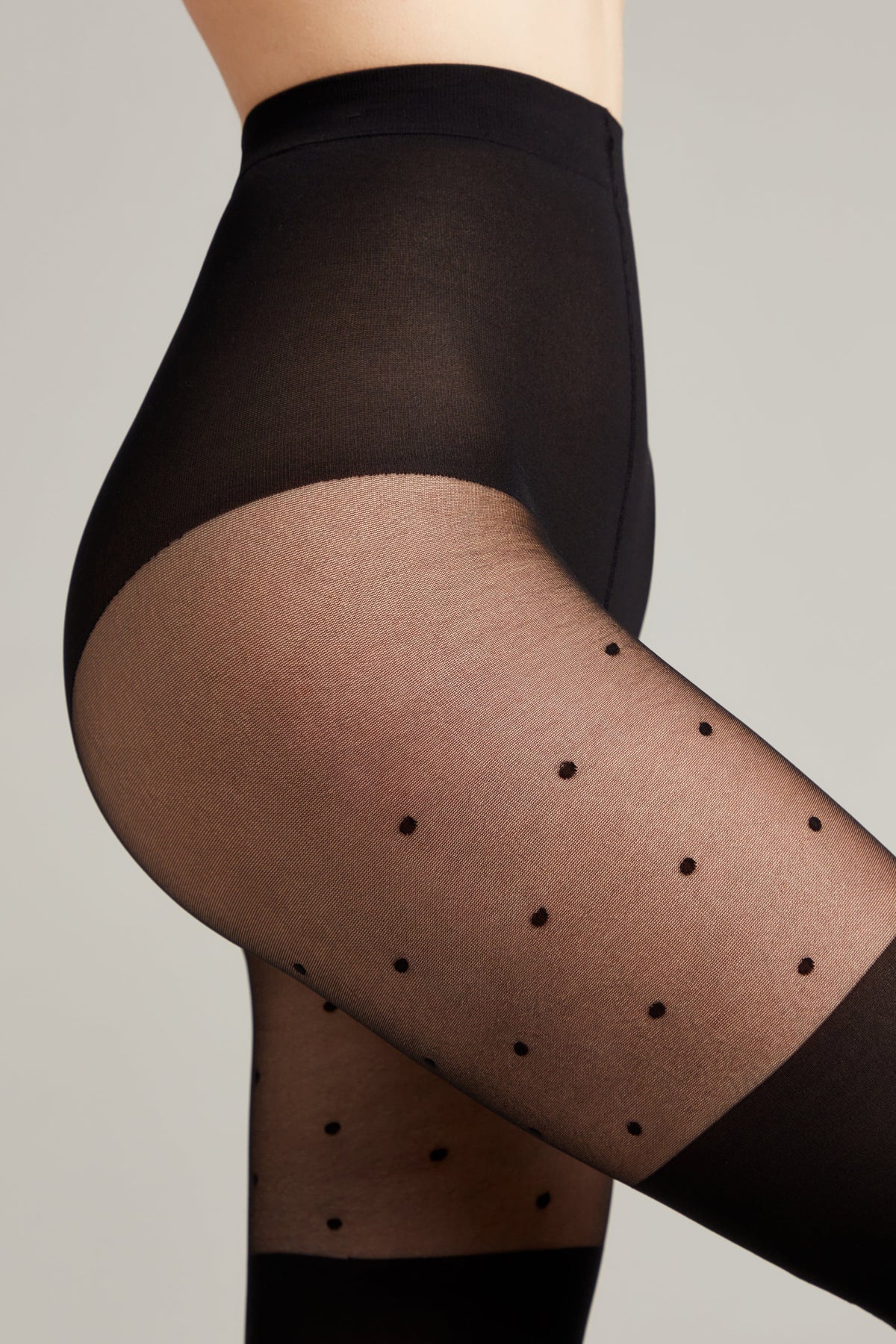 Close-up side view of a person wearing black sheer tights with a small polka dot pattern on a light grey background.