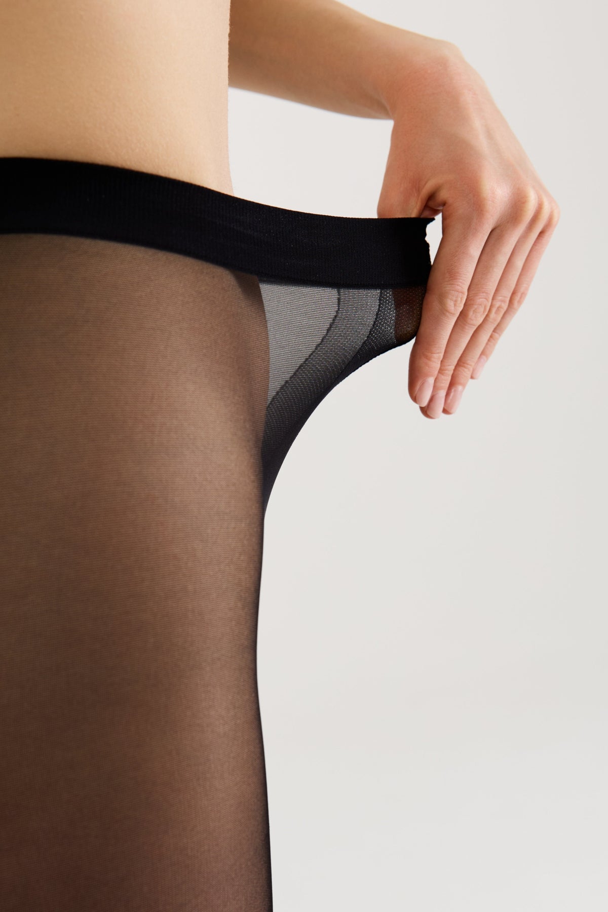 A close-up view of a person wearing sheer black tights, holding the elastic waistband with one hand against a plain background.