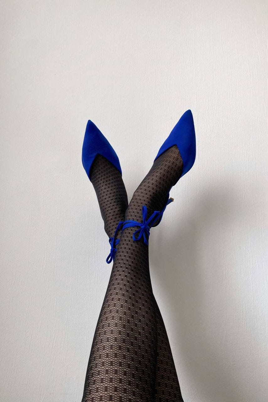 A pair of pointed-toe bright blue heels with ankle ties worn with black patterned tights against a plain white wall.