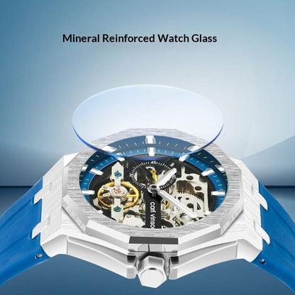 A skeletonized wristwatch with a silver octagonal bezel, blue dial, and blue rubber strap is displayed with its round mineral reinforced glass hovering slightly above the watch face.