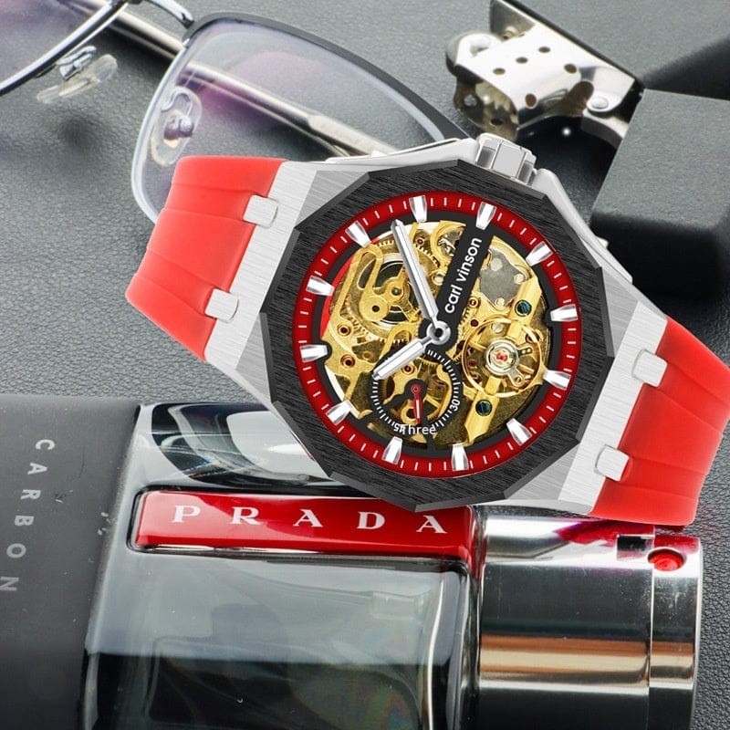 A skeleton watch featuring a silver octagonal case, a red and black bezel, and a red rubber strap rests on top of a black bottle.