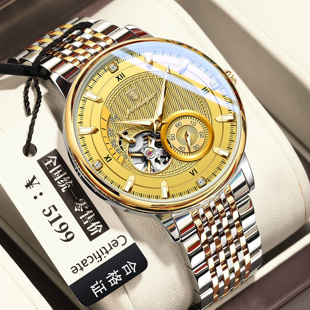 A round stainless steel analog watch features a gold-toned face with a textured pattern, visible mechanical movement, silver and gold-toned metal link band, and an attached white product tag.