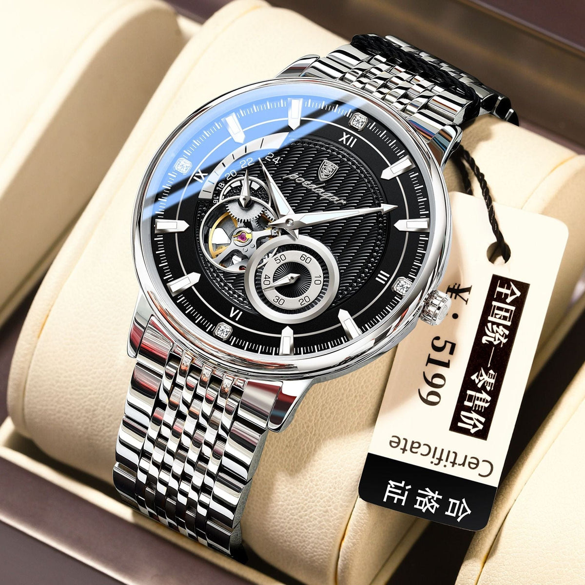 A silver-toned men's analog watch with a round black dial, intricate gear detailing, a stainless steel link bracelet, and a white price tag attached, resting on a beige display cushion.