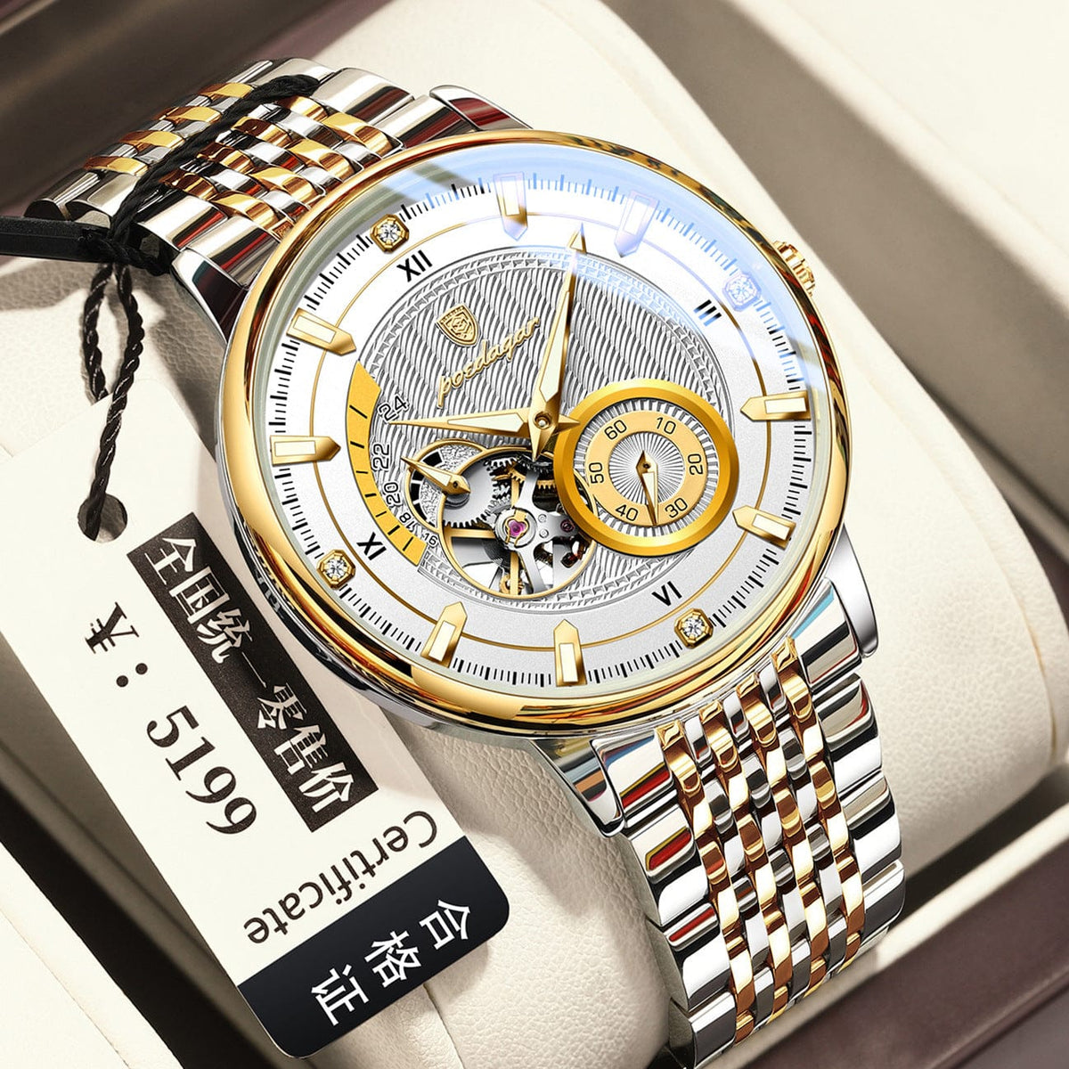 A silver and gold-tone analog watch featuring a round face with an intricate metallic pattern, a skeleton cutaway, a circular sub-dial, and a two-toned stainless steel link bracelet resting in a display box with an attached tag.